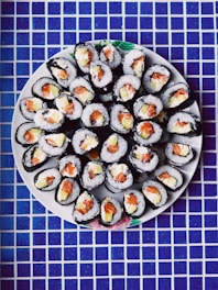 An arrangement of sushi rolls on a round white plate. The rolls are neatly placed, showcasing fillings of salmon, rice, and possibly avocado. The background features a blue and white checkered tile surface.