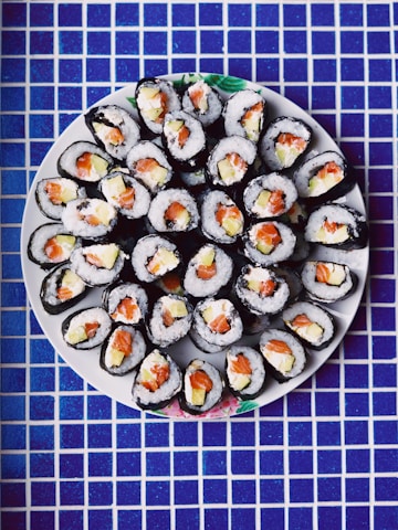 An arrangement of sushi rolls on a round white plate. The rolls are neatly placed, showcasing fillings of salmon, rice, and possibly avocado. The background features a blue and white checkered tile surface.