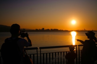 Silhouette of a man with a camera, a child, and a woman pointing at the sunset over a body of water. The skyline in the distance is dark against the vibrant orange and yellow hues of the sky.