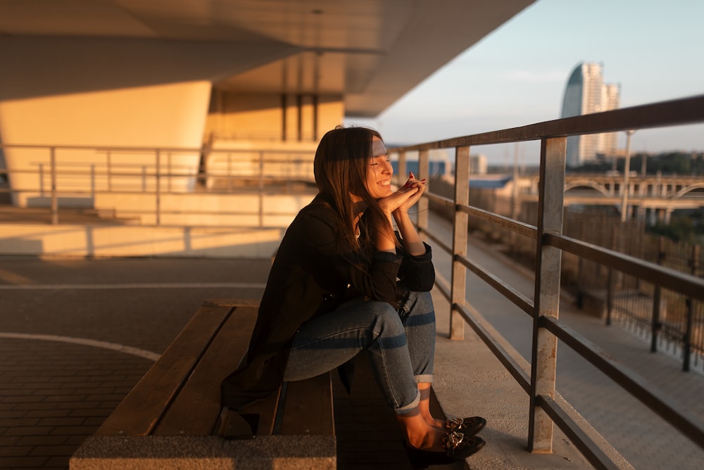 woman in black cardigan and blue jeans sitting on bench by rail