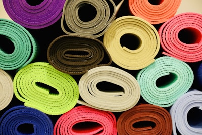 A collection of neatly rolled mats or fabric, each rolled up tightly and arranged in a grid. The rolls display a variety of vibrant colors, including purple, pink, green, blue, and brown. The texture of the material appears soft and slightly fuzzy.