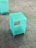 A small, translucent, blue-green cube-shaped acrylic stool or display stand is placed on a tiled floor. The surface has a smooth finish, giving it a modern and minimalist appearance.