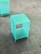 A small, translucent, blue-green cube-shaped acrylic stool or display stand is placed on a tiled floor. The surface has a smooth finish, giving it a modern and minimalist appearance.