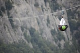 A person wearing a green helmet is zip-lining through a mountainous area. The background consists of rugged, gray rock formations with patches of greenery.