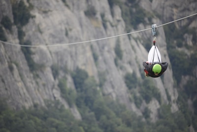 A person wearing a green helmet is zip-lining through a mountainous area. The background consists of rugged, gray rock formations with patches of greenery.
