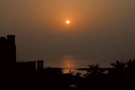 A serene sunset over the ocean, with the sun casting a warm glow across the water. Silhouettes of palm trees and architectural structures create a peaceful silhouette against the vibrant sky.