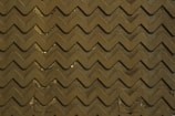 A zigzag patterned surface, appearing to be a textured mat or flooring. The pattern is uniformly repeated across the surface, with a few small areas of dirt and debris visible between the raised sections.