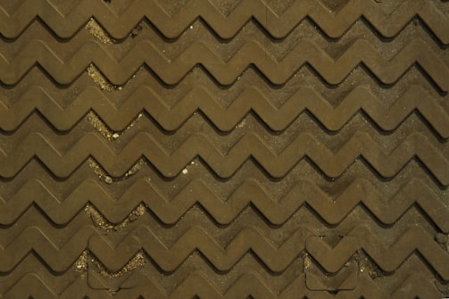 A zigzag patterned surface, appearing to be a textured mat or flooring. The pattern is uniformly repeated across the surface, with a few small areas of dirt and debris visible between the raised sections.