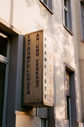A wooden sign with text in German hangs on the wall of a building. It appears weathered, with shadows cast on the wall, and includes details like numbers and words in blue and orange.