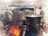 A professional kitchen with chefs at work wearing white uniforms and tall hats. Large pots are steaming, and the stove is actively in use with visible flames. The busy atmosphere suggests the preparation of a meal with various equipment visible in the background.