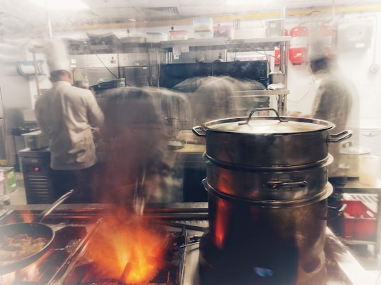 A professional kitchen with chefs at work wearing white uniforms and tall hats. Large pots are steaming, and the stove is actively in use with visible flames. The busy atmosphere suggests the preparation of a meal with various equipment visible in the background.