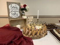 A dessert table is elegantly arranged with a wooden crate sign that reads 'love is sweet, please take a treat' in decorative script. The table is adorned with a burgundy cloth, a white candle, and a glass vase holding red and white roses. A variety of desserts are neatly displayed, including churros in cups, cheesecake, and chocolate drizzled pastries. Small cards are placed next to each dessert to label them.