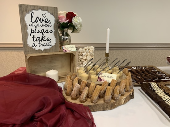 A dessert table is elegantly arranged with a wooden crate sign that reads 'love is sweet, please take a treat' in decorative script. The table is adorned with a burgundy cloth, a white candle, and a glass vase holding red and white roses. A variety of desserts are neatly displayed, including churros in cups, cheesecake, and chocolate drizzled pastries. Small cards are placed next to each dessert to label them.