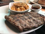 A plate with grilled ribs seasoned with pepper and a portion of creamy macaroni and cheese in a small white dish. A small cup of barbecue sauce is also present on the plate.