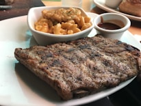A plate with grilled ribs seasoned with pepper and a portion of creamy macaroni and cheese in a small white dish. A small cup of barbecue sauce is also present on the plate.