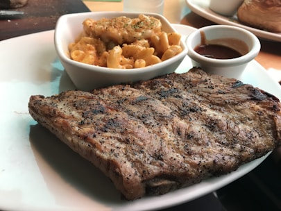 A plate with grilled ribs seasoned with pepper and a portion of creamy macaroni and cheese in a small white dish. A small cup of barbecue sauce is also present on the plate.
