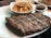 A plate with grilled ribs seasoned with pepper and a portion of creamy macaroni and cheese in a small white dish. A small cup of barbecue sauce is also present on the plate.