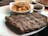 A plate with grilled ribs seasoned with pepper and a portion of creamy macaroni and cheese in a small white dish. A small cup of barbecue sauce is also present on the plate.