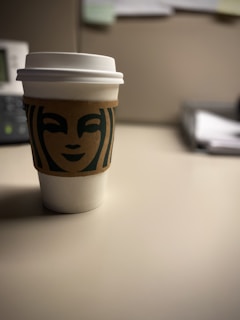 A paper coffee cup with a white lid and a brown sleeve featuring a green logo is placed on a neutral-colored table. The background is slightly out of focus, showing the corner of an office desk setup with an organized stack of papers.