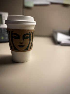 A paper coffee cup with a white lid and a brown sleeve featuring a green logo is placed on a neutral-colored table. The background is slightly out of focus, showing the corner of an office desk setup with an organized stack of papers.