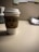 A paper coffee cup with a white lid and a brown sleeve featuring a green logo is placed on a neutral-colored table. The background is slightly out of focus, showing the corner of an office desk setup with an organized stack of papers.