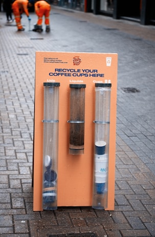 A colorful recycling station with three transparent tubes for collecting coffee cup lids, liquids, and cups against a street backdrop. Two individuals wearing orange work uniforms are in the background, engaged in street maintenance.