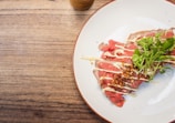 A close-up of a plate with thinly sliced raw meat, likely beef, garnished with creamy sauce and topped with crispy fried onions and fresh green herbs. The plate is placed on a wooden surface with a cup partially visible near the top edge.