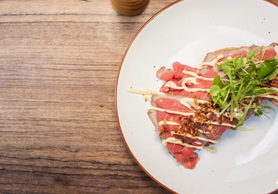A close-up of a plate with thinly sliced raw meat, likely beef, garnished with creamy sauce and topped with crispy fried onions and fresh green herbs. The plate is placed on a wooden surface with a cup partially visible near the top edge.