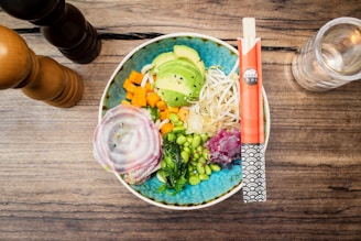 A bowl of vibrant, fresh vegetables arranged artfully on a wooden table. Ingredients include avocado slices, bean sprouts, diced carrots, edamame beans, sliced radish, seaweed, and purple cabbage, served with a pair of colorful chopsticks. Nearby are a wooden pepper grinder and a glass of water.