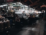 A bustling indoor market with numerous stalls displaying a variety of goods such as packaged foods, snacks, and souvenirs. The lighting is dim, but individual bulbs illuminate the items for sale. Shoppers and vendors are interacting, creating a lively atmosphere.