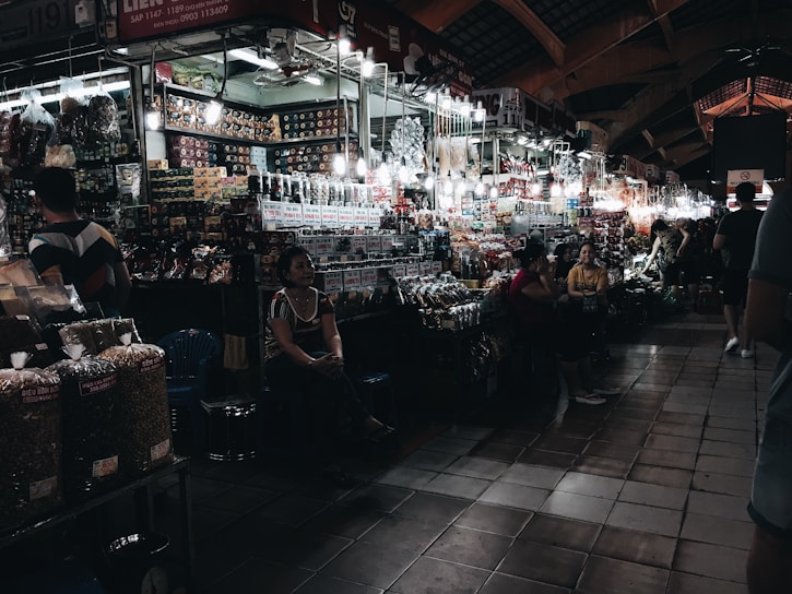 A bustling indoor market with numerous stalls displaying a variety of goods such as packaged foods, snacks, and souvenirs. The lighting is dim, but individual bulbs illuminate the items for sale. Shoppers and vendors are interacting, creating a lively atmosphere.