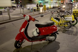 A red scooter is parked on a city street next to several bicycles, including yellow and black electric bikes. The background features blurred motion of vehicles and a tram, suggesting nighttime or dusk in an urban setting.