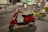 A red scooter is parked on a city street next to several bicycles, including yellow and black electric bikes. The background features blurred motion of vehicles and a tram, suggesting nighttime or dusk in an urban setting.