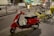 A red scooter is parked on a city street next to several bicycles, including yellow and black electric bikes. The background features blurred motion of vehicles and a tram, suggesting nighttime or dusk in an urban setting.