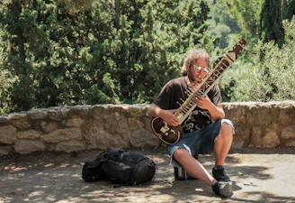 A person with long hair and sunglasses sits on a low rock wall outdoors, playing a sitar. Nearby, a black bag rests on the ground. Tall, lush green trees provide a natural background.