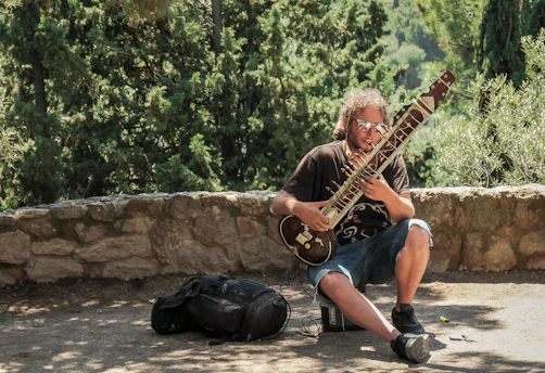 A person with long hair and sunglasses sits on a low rock wall outdoors, playing a sitar. Nearby, a black bag rests on the ground. Tall, lush green trees provide a natural background.