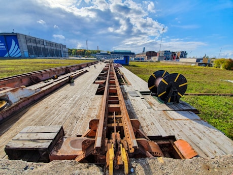 A wooden platform on a railway track extends into the distance under a partly cloudy sky. Large industrial buildings are visible in the background. On the right, there is a large circular spool or reel, possibly for cable or hose storage. The surrounding area is grassy with some scattered construction elements.