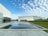 A modern architecture scene featuring sleek, white buildings with clean lines and expansive glass walls. In the foreground, a reflective pool mirrors the sky and surrounding structures. The sky is filled with a pattern of soft, white clouds against a blue backdrop, adding to the serene atmosphere. The landscape includes a neatly maintained grass area.