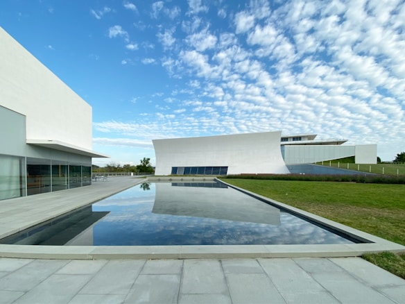 A modern architecture scene featuring sleek, white buildings with clean lines and expansive glass walls. In the foreground, a reflective pool mirrors the sky and surrounding structures. The sky is filled with a pattern of soft, white clouds against a blue backdrop, adding to the serene atmosphere. The landscape includes a neatly maintained grass area.