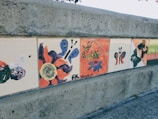 A series of hand-painted tiles mounted on a concrete wall featuring various abstract designs, including butterflies, abstract shapes, and bold colors. One tile reads 'The Attics' with some artistic brush strokes.