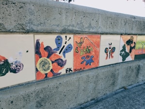 A series of hand-painted tiles mounted on a concrete wall featuring various abstract designs, including butterflies, abstract shapes, and bold colors. One tile reads 'The Attics' with some artistic brush strokes.