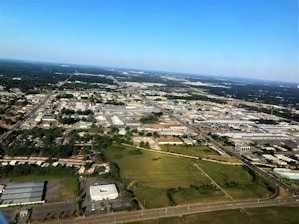 Aerial view of an urban area with a mix of industrial buildings, commercial spaces, and residential neighborhoods. A network of roads and streets is visible, intersecting the landscape. In the foreground, there is an open green space, while in the distance, the horizon is visible with a clear blue sky above.