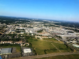 Aerial view of an urban area with a mix of industrial buildings, commercial spaces, and residential neighborhoods. A network of roads and streets is visible, intersecting the landscape. In the foreground, there is an open green space, while in the distance, the horizon is visible with a clear blue sky above.