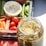 A glass jar filled with sliced pickles sits in the foreground, open with liquid visible inside. In the background, a platter containing a variety of fresh vegetables such as green beans, baby carrots, cherry tomatoes, and celery can be seen. A cup of dip is also visible near the vegetables, suggesting a snack or appetizer setting. The image is well-lit with daylight, highlighting the freshness and vibrant colors of the produce.