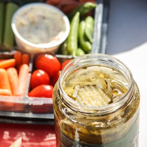 A glass jar filled with sliced pickles sits in the foreground, open with liquid visible inside. In the background, a platter containing a variety of fresh vegetables such as green beans, baby carrots, cherry tomatoes, and celery can be seen. A cup of dip is also visible near the vegetables, suggesting a snack or appetizer setting. The image is well-lit with daylight, highlighting the freshness and vibrant colors of the produce.