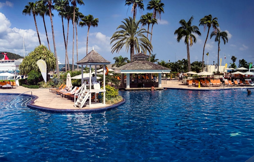 A tropical resort with a large swimming pool surrounded by palm trees and lounge chairs. There is a shaded cabana area near the pool and people are relaxing by the water. A cruise ship is in the background against a clear blue sky.