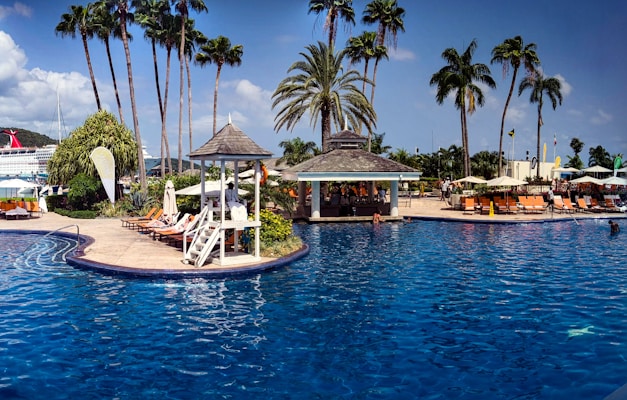 A tropical resort with a large swimming pool surrounded by palm trees and lounge chairs. There is a shaded cabana area near the pool and people are relaxing by the water. A cruise ship is in the background against a clear blue sky.