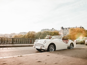 A classic white convertible car is parked on a bridge with elegant buildings and lush trees in the background. The car is decorated with a bouquet of flowers on the hood, suggesting a celebratory or wedding theme. The scene is bathed in a warm, golden sunlight creating a romantic atmosphere.