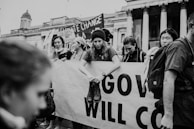 A group of people is participating in a protest, holding a large banner with partial visible text. One person is sitting on the banner, wearing a beanie and holding boots. The scene includes signs about climate change. The architecture of the building in the background suggests a public space.
