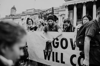 A group of people is participating in a protest, holding a large banner with partial visible text. One person is sitting on the banner, wearing a beanie and holding boots. The scene includes signs about climate change. The architecture of the building in the background suggests a public space.